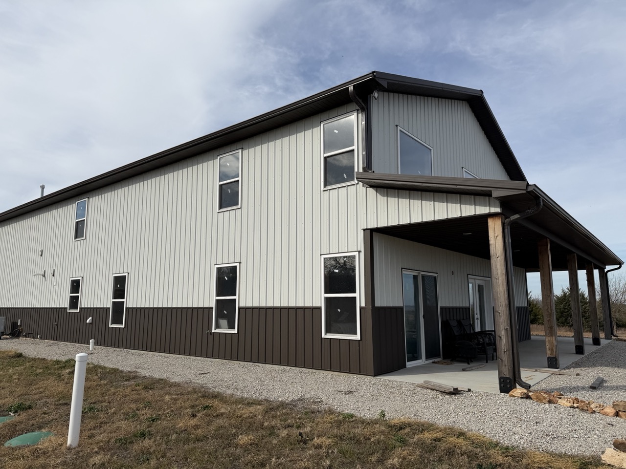Post-frame barndominium exterior with light metal siding, dark lower metal panels, multiple residential windows, and a covered porch supported by timber posts.