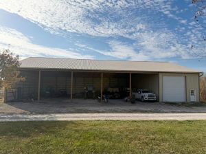 Post-frame building featuring a wide open lean-to section supported by wood posts, metal siding and roof, enclosed garage bay, and stored vehicles and equipment on a gravel drive.