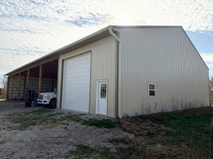 Post-frame garage building with light metal siding, tall overhead garage door, side entry door, and an attached open lean-to bay used for vehicle storage on a gravel base.