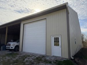 Post-frame garage building with light metal siding, a tall white overhead garage door, side entry door, and an open lean-to bay housing a pickup truck on a gravel surface.